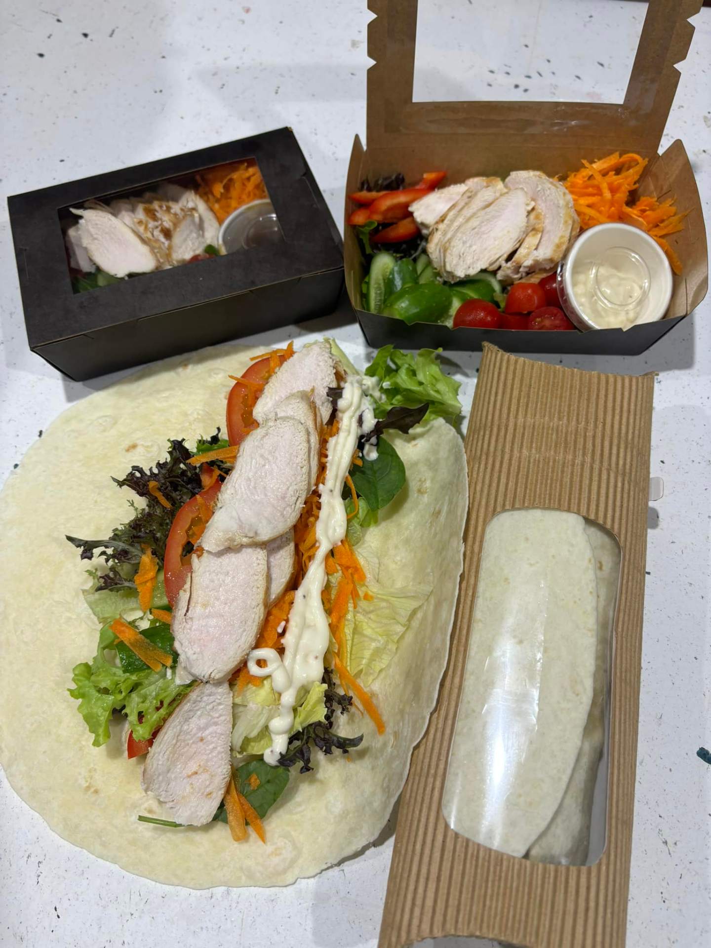 Chicken and salad takeaway boxes with carrot, cucumber and tomato, alongside a fresh chicken salad wrap